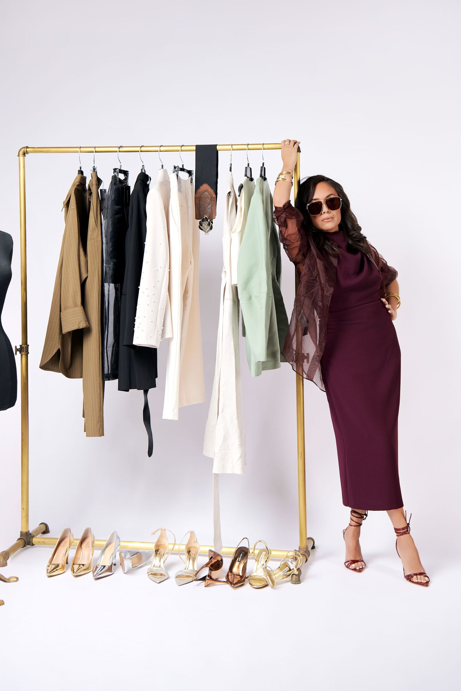 Stylish clothing rack with neutral garments and gold metal frame next to assorted heels and burgundy dress outfit.