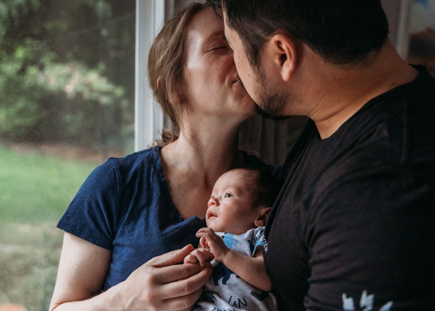 A couple shares a tender moment while holding their newborn baby in a navy onesie by a window.