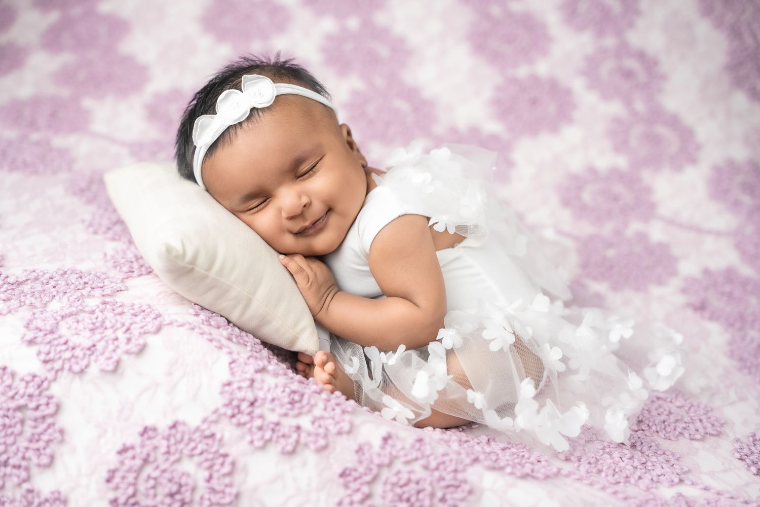 A sleeping baby in white lace outfit lies on soft pink damask fabric background with delicate floral patterns.