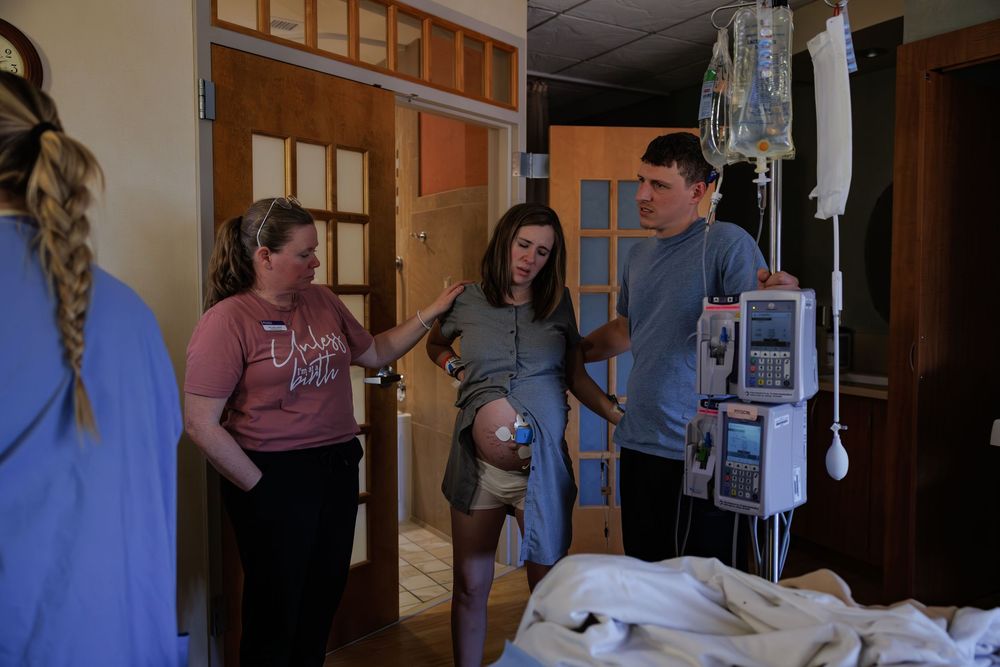 People gathered around a hospital bed with medical equipment and monitors visible in the background.