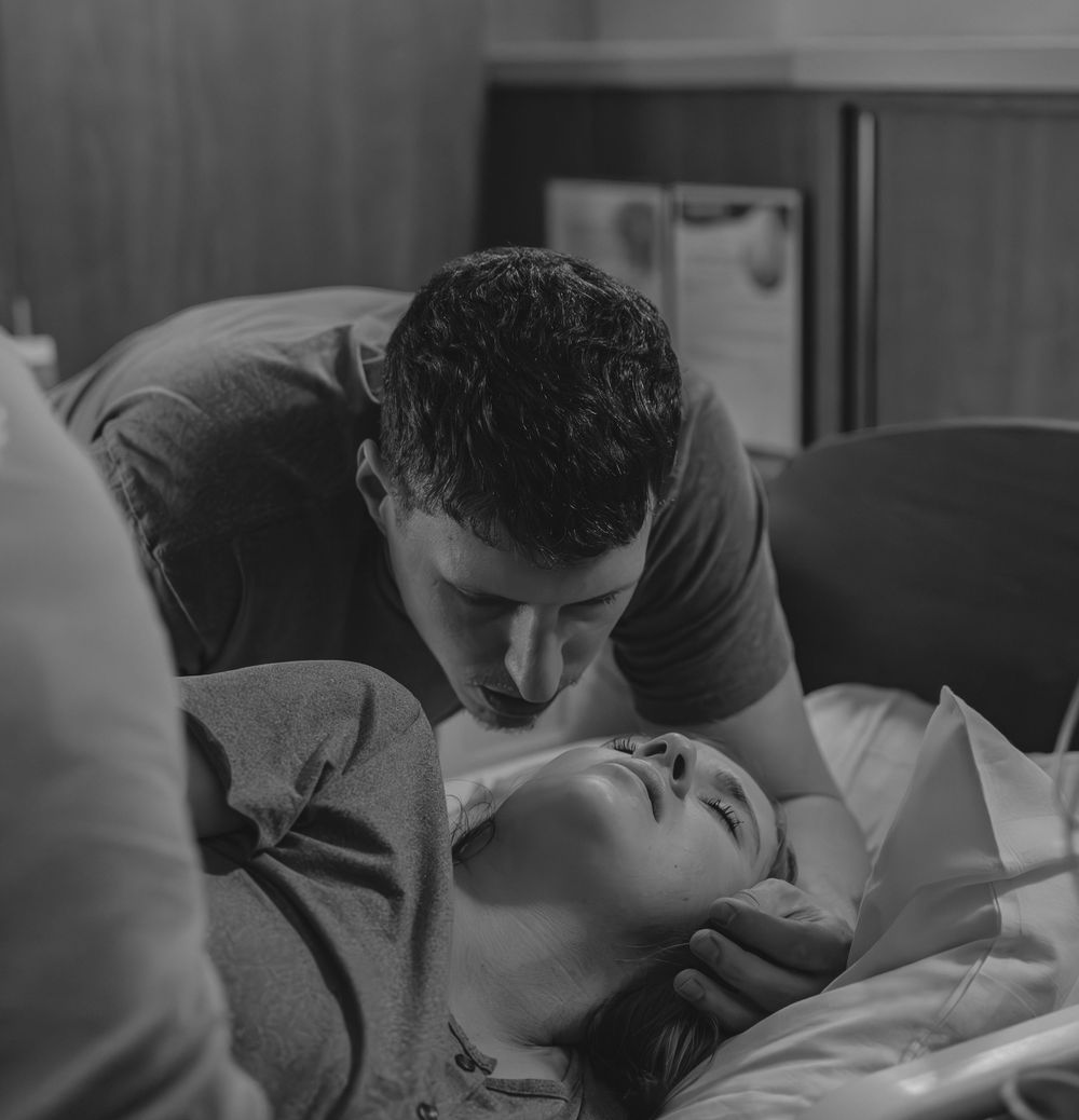 A tender black and white scene showing an intimate moment between two people, with one leaning over the other on a bed.