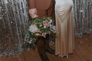 A couple in wedding attire poses against a silver sequin backdrop, with the bride holding a pastel floral bouquet.