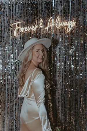 A bride in a silk dress and white hat poses in front of a sparkly backdrop with neon signage.