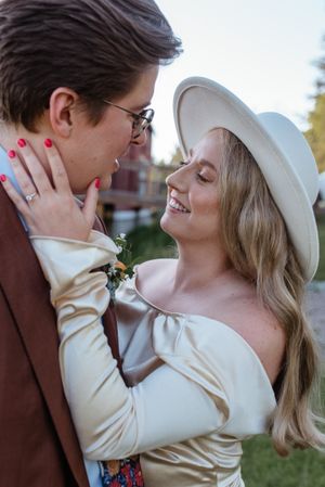 A couple shares an intimate moment outdoors, with one wearing a brown suit and the other in white.