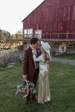 A couple in wedding attire poses romantically in front of a rustic red barn at sunset.