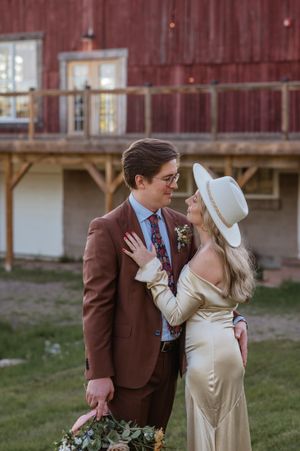 A couple in vintage wedding attire poses intimately against a rustic red barn backdrop.
