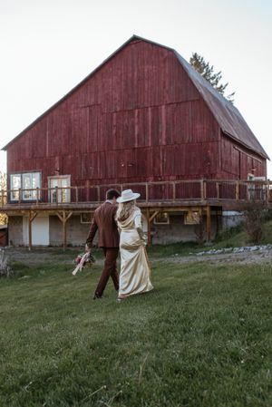 A couple walking hand in hand in front of a classic red barn during golden hour.