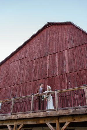 A couple stands on the balcony of a large red barn during their wedding photoshoot.
