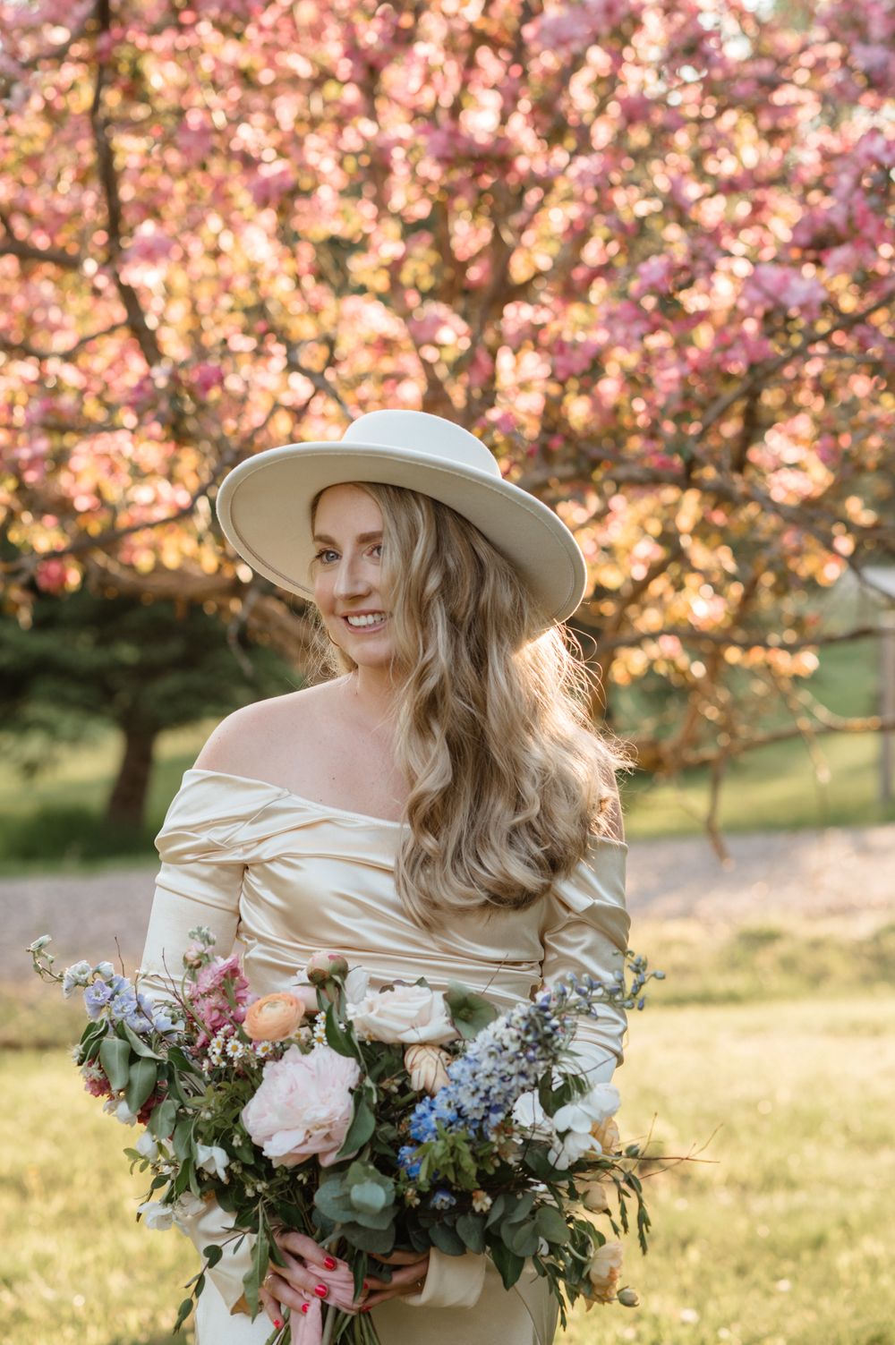 Spring portrait under cherry blossoms with cream dress and wide brim hat holding floral bouquet.