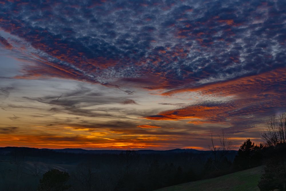 Colorful dramatic sunset with orange and blue clouds over a rolling forested hillside.