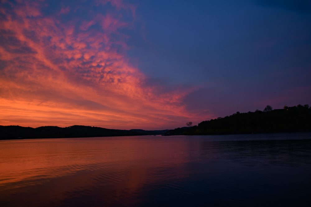 Dramatic red and orange sunset reflecting over a calm lake with silhouetted tree line.