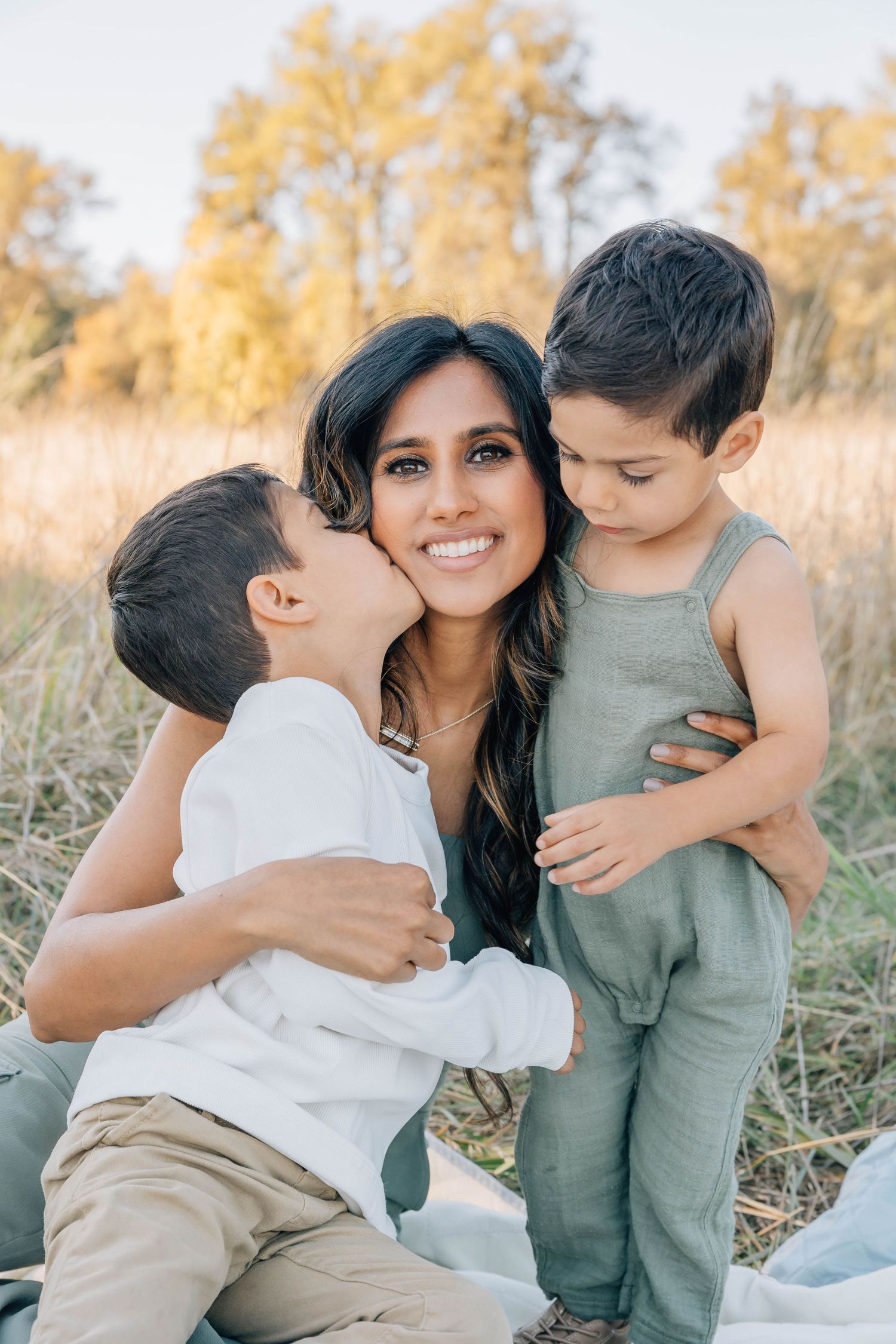 Smiling mother with two young sons sharing a kiss outdoors in golden field at sunset.