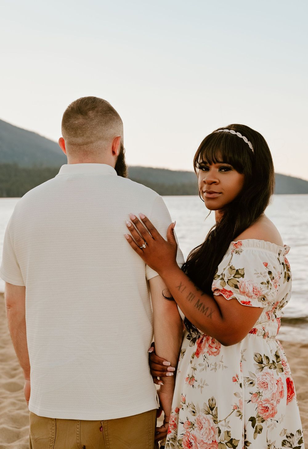 A couple stands together by a serene lake at sunset, with one wearing a floral off-shoulder dress and the other in white.