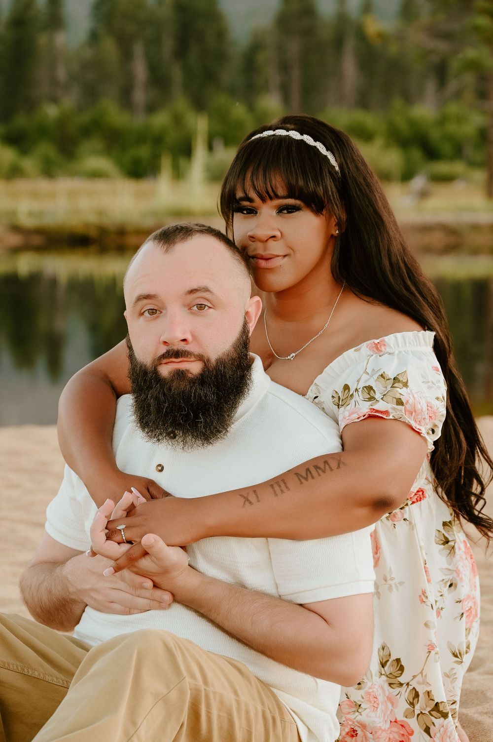 Romantic couple embraces by a lake during sunset, with her wearing a floral dress and him in white with a full beard.