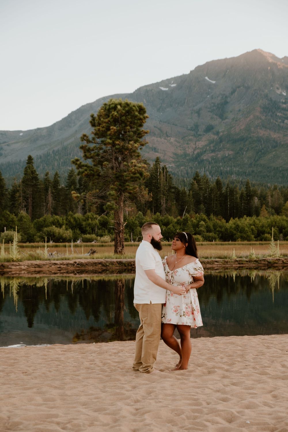 A couple embraces during a lakeside photo session with mountains and evergreen trees in the background at sunset.