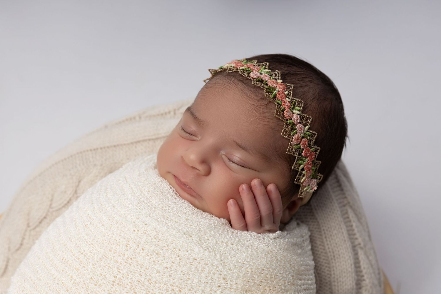 A sleeping newborn wrapped in a peach blanket lies on lace fabric with cherry blossoms beside them on wooden background.