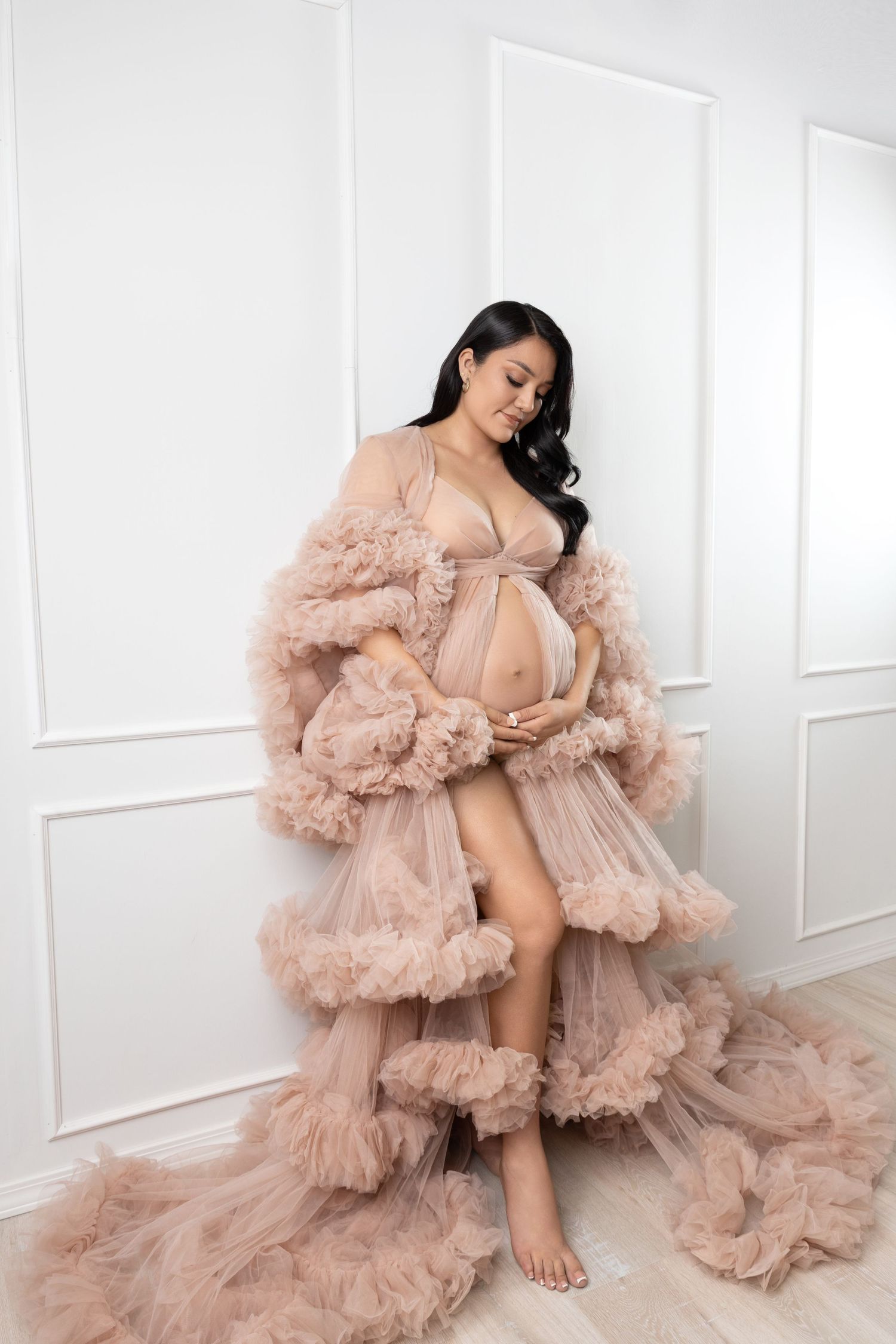 Expectant woman in flowing mauve gown and floral crown poses elegantly against neutral backdrop in maternity photo.
