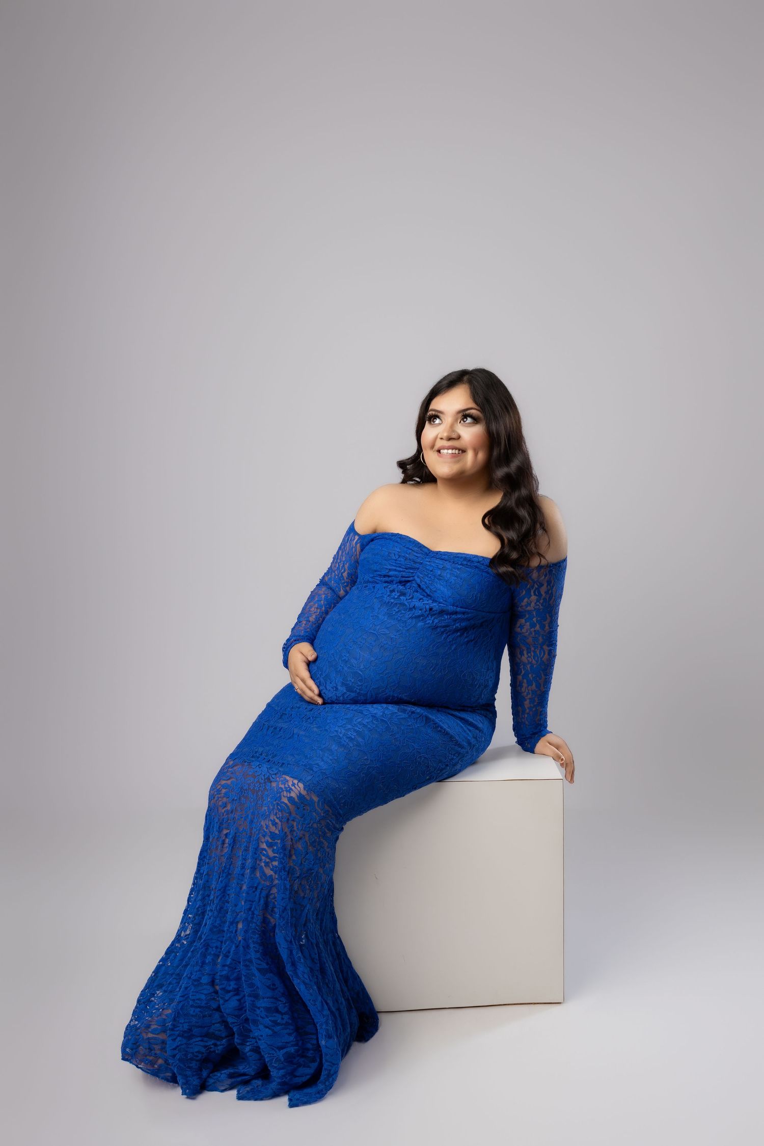 Expectant woman in flowing mauve gown and floral crown poses elegantly against neutral backdrop in maternity photo.