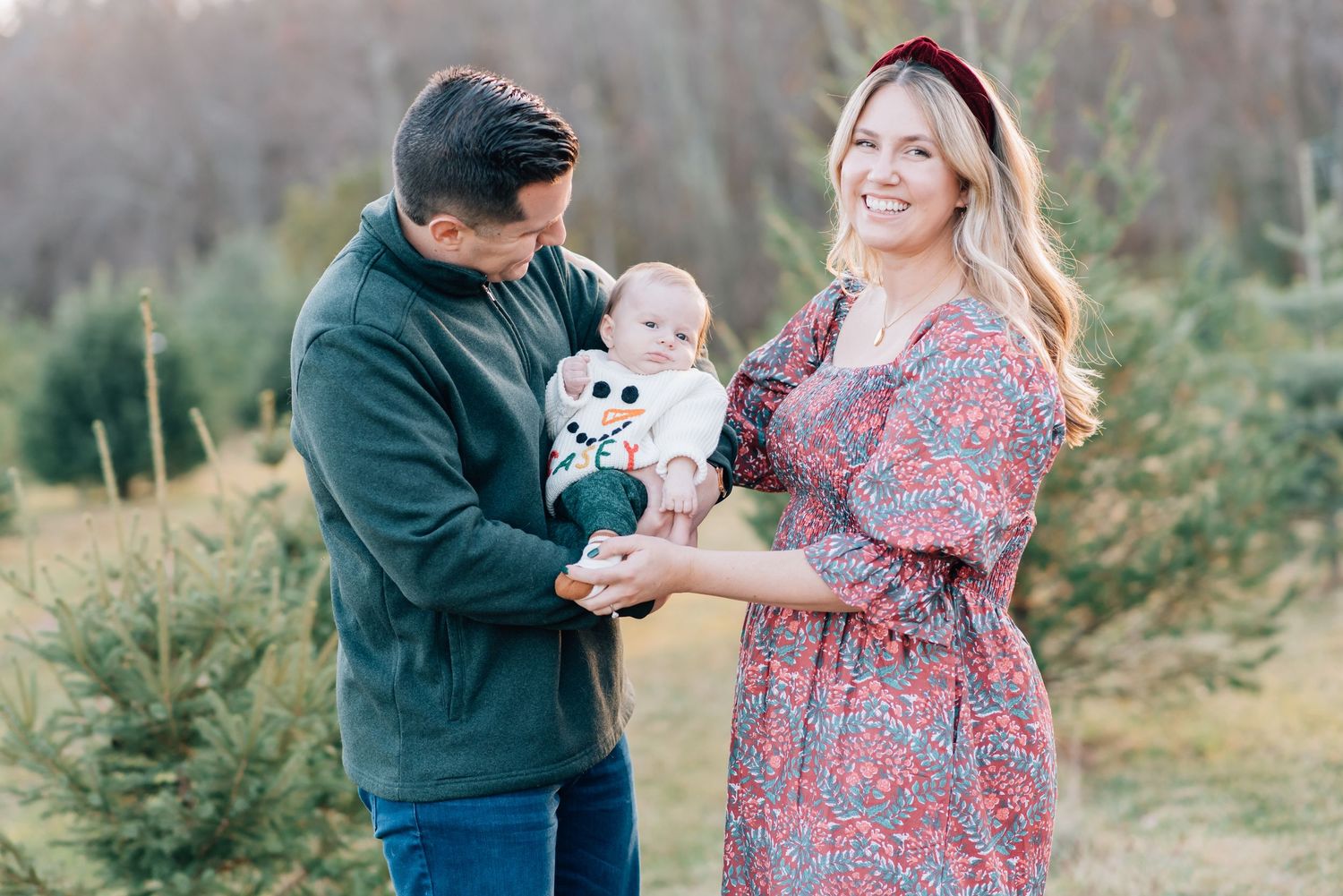 A man in a green sweater and his wife holding their child while smiling and posing for a holiday Christmas card photo.