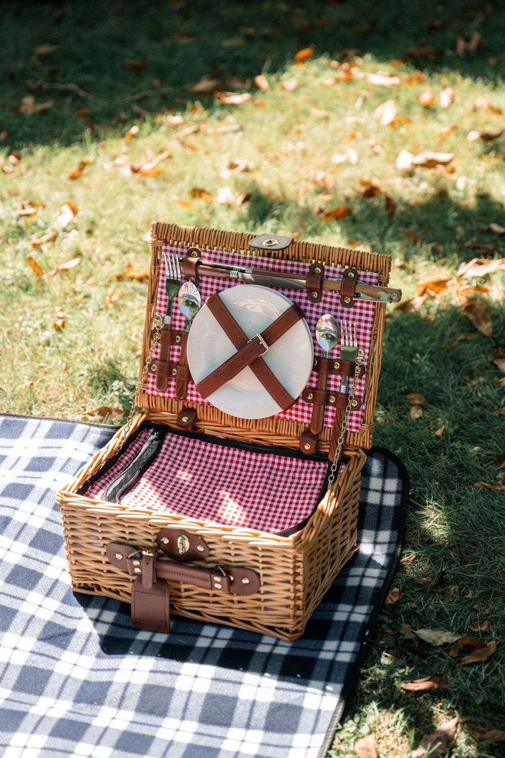 Vintage wicker picnic basket with red gingham lining and plates sits on a checkered blanket in dappled sunlight.