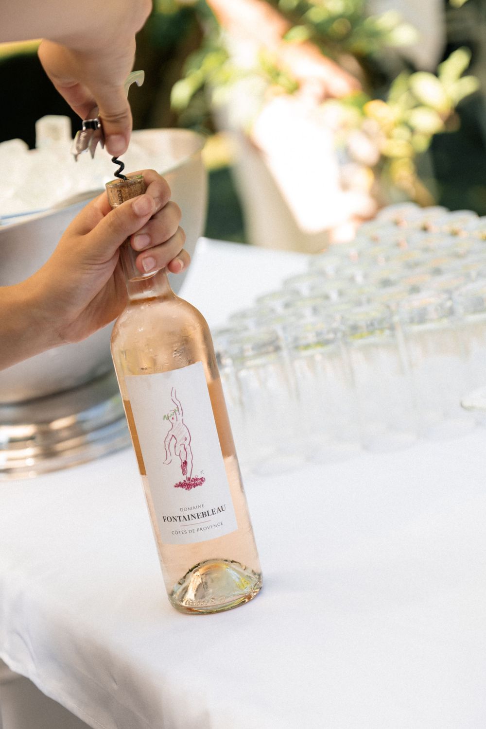 Hands opening a bottle of rose wine next to empty glasses on a white tablecloth in outdoor setting.