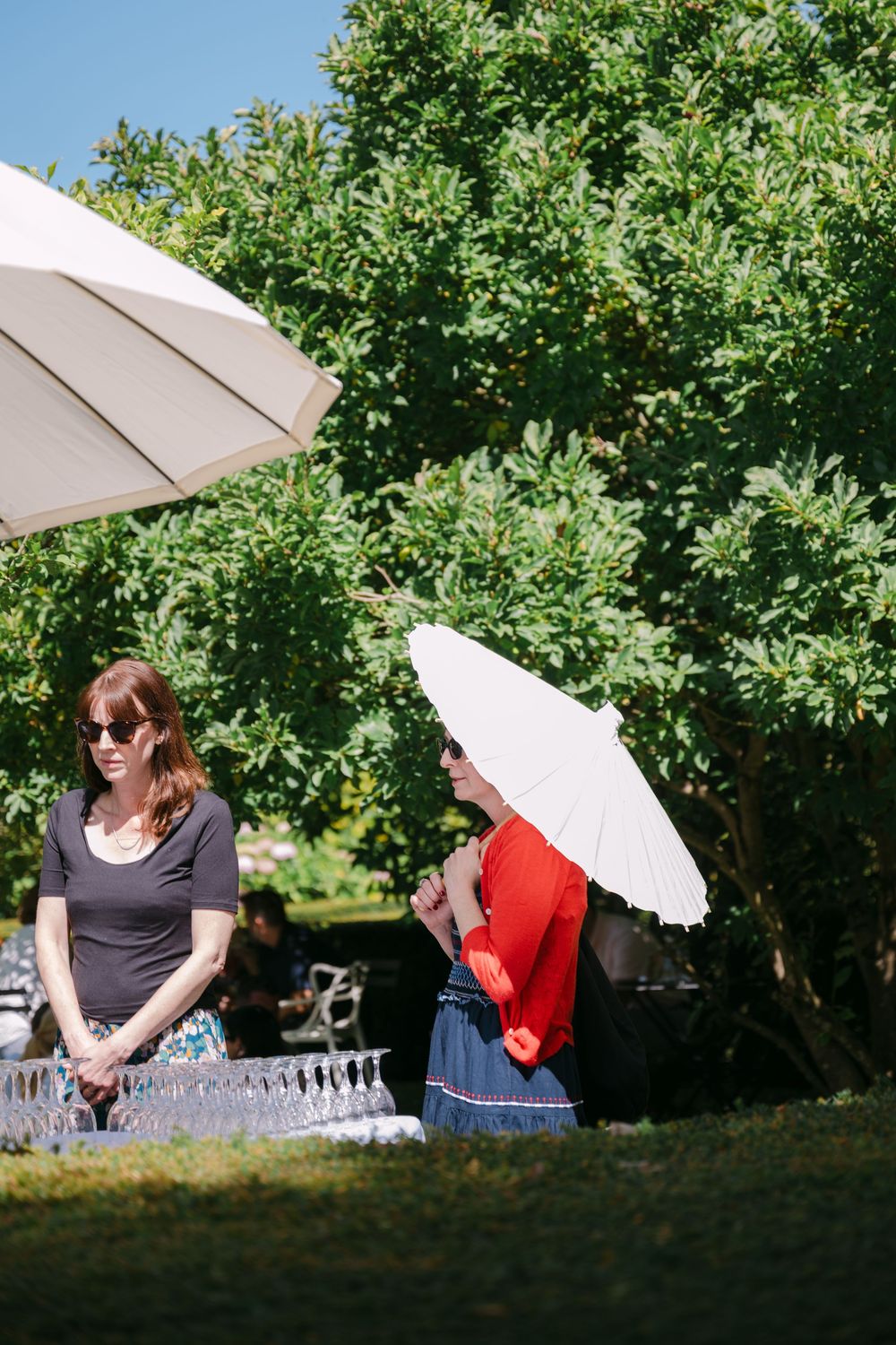 People relaxing with white parasols under green trees on a sunny afternoon.