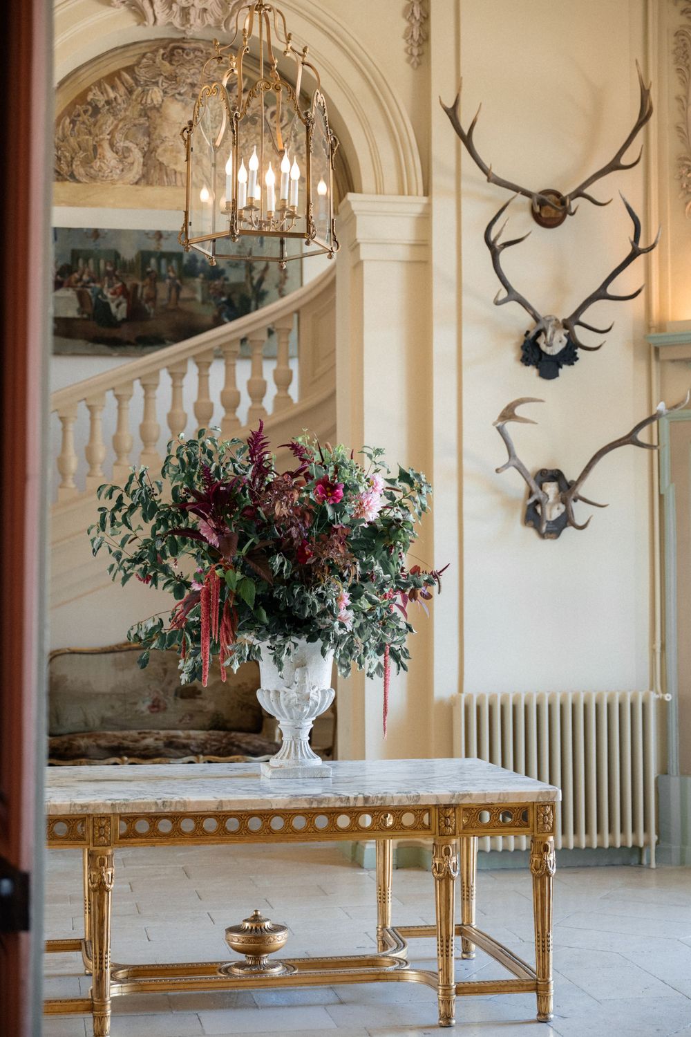 Elegant hallway with antique decor featuring mounted antlers, ornate tables, and floral arrangements in urns.
