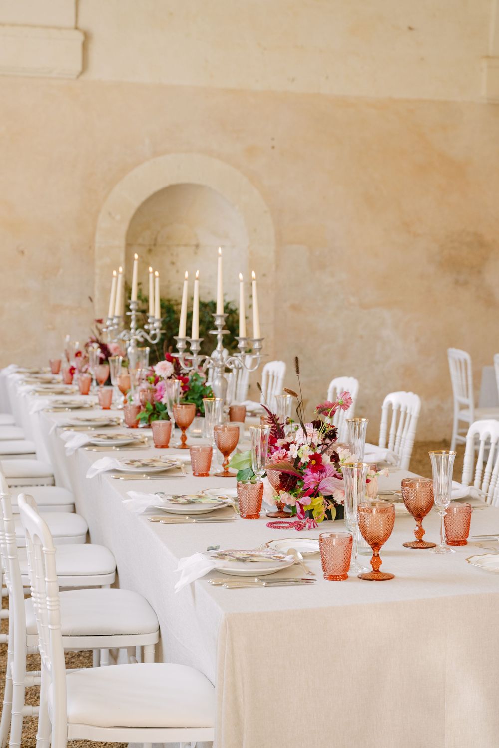 Elegant wedding reception table setting with white chairs, pink glassware, and white candles in a rustic venue.