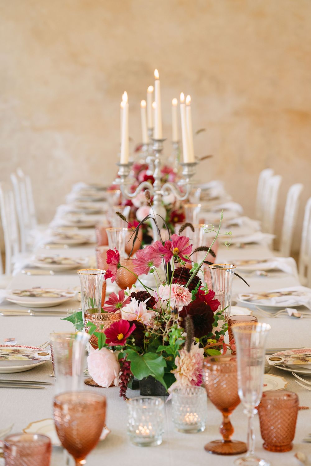 Long dining table with romantic candlelight, pink glassware, and vibrant floral arrangements in burgundy and blush tones.