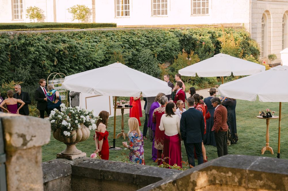 Outdoor garden party with white canopy tents and guests gathered on a stone terrace near a historic building.