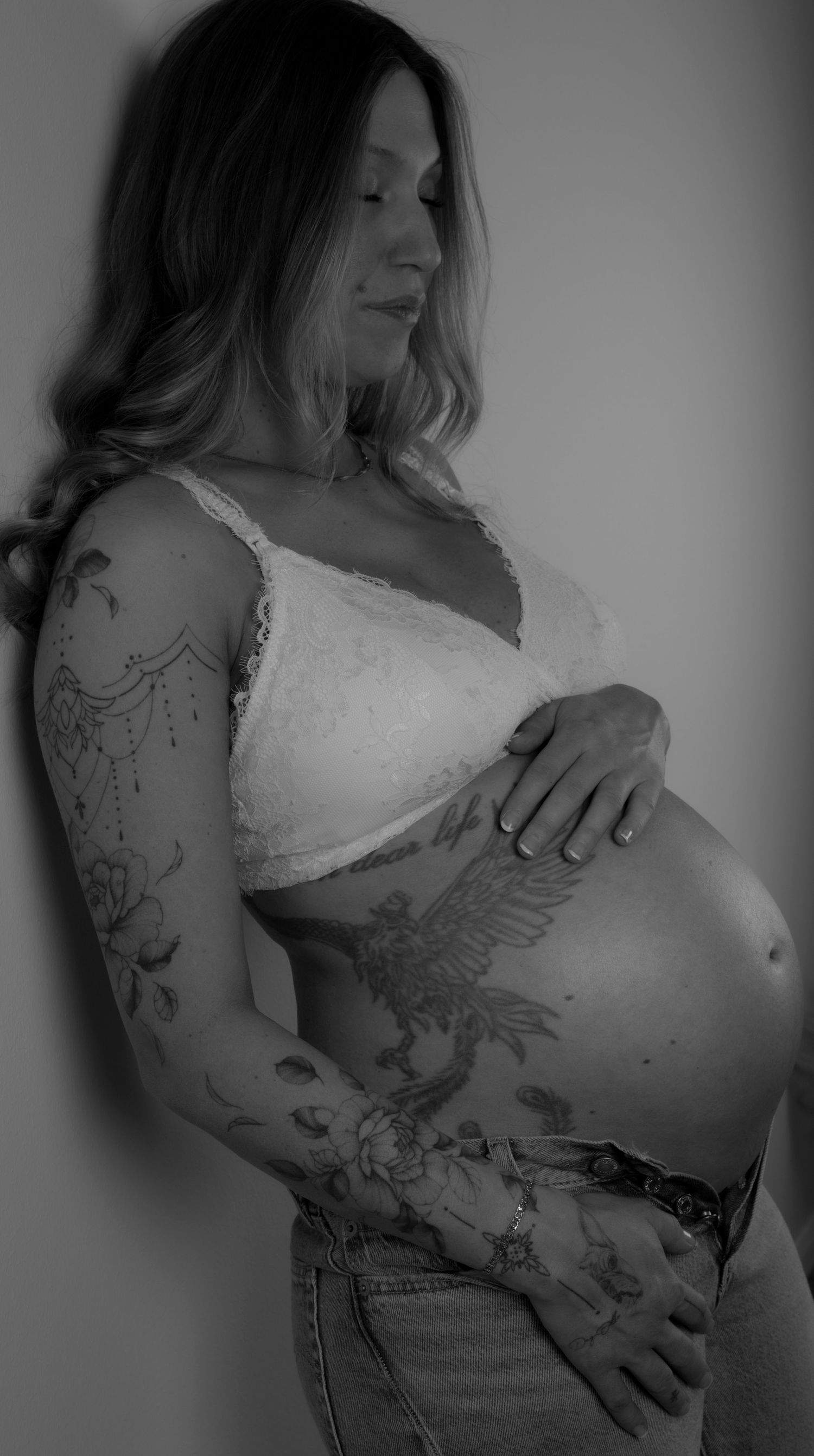 fotografering av en gravid kvinna som står i en topp och ett par jeans och håller handen på sin gravidmage. bilden är svartvit.