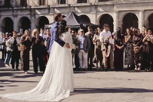 Novios de pie en plaza histórica rodeados de invitados, con vestido blanco y ramo floral, la Plaza España, Vitoria-Gasteiz