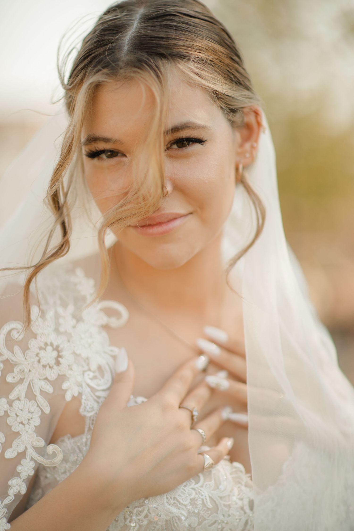 A bride wearing a delicate lace veil smiles warmly while showing her wedding ring in soft natural lighting.