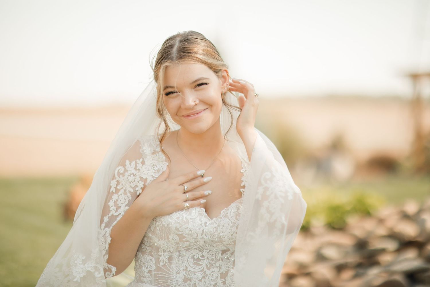 A bride in a detailed lace wedding dress and veil poses outdoors in warm sunlight against a natural landscape.