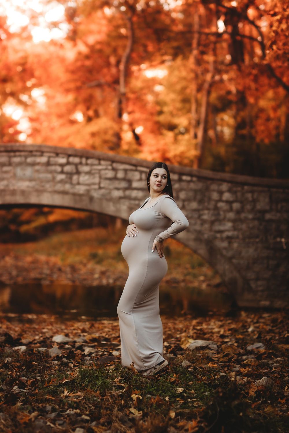 Pregnant woman in form-fitting beige dress poses near stone bridge surrounded by vibrant red autumn foliage.