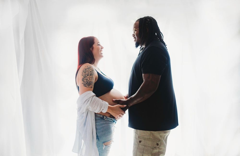 Pregnant tattooed woman and partner holding hands facing each other against white backdrop.