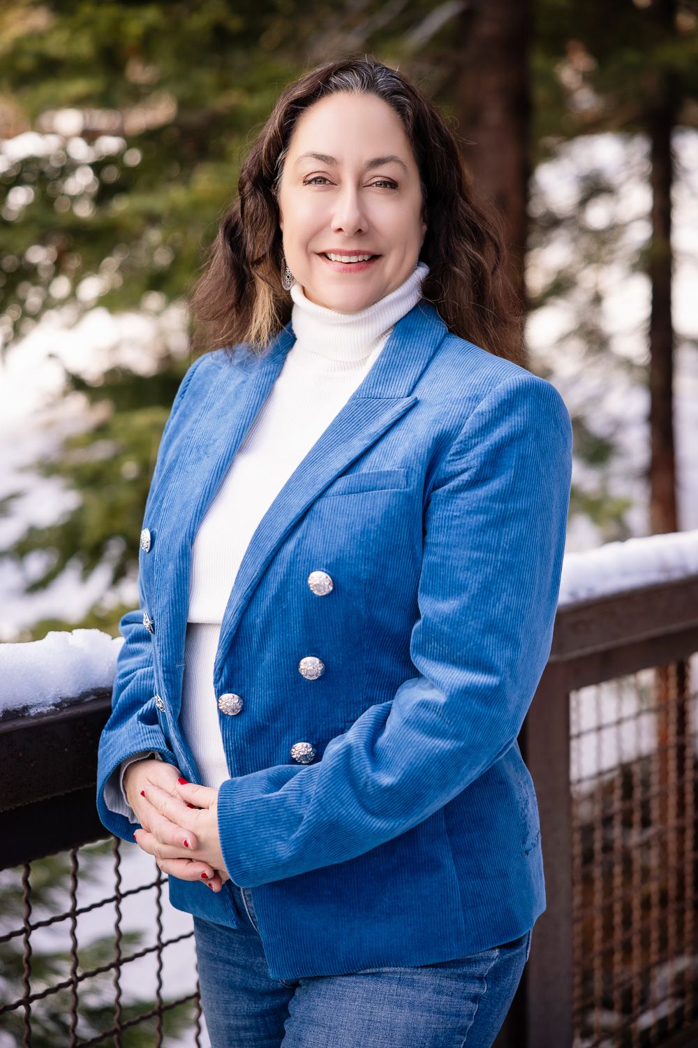 A person in a bright blue blazer and white top poses outdoors against a snowy winter backdrop with evergreen trees.