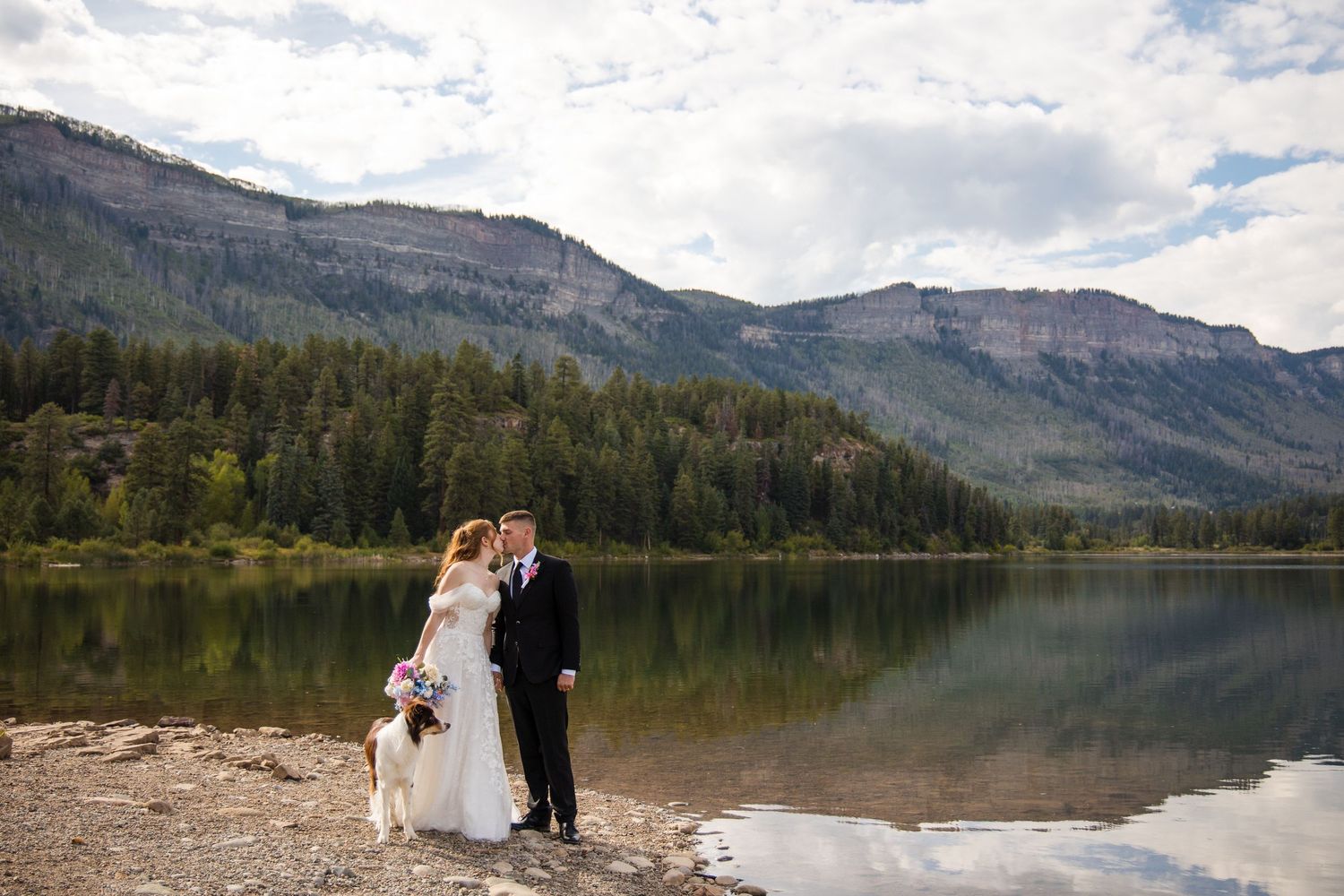 A couple stands on a serene lakeshore with mountain views and forest backdrop during a wedding ceremony.