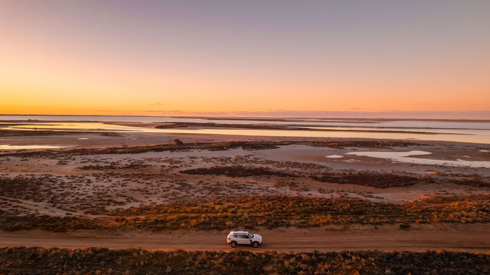 White SUV driving along coastal dirt road at sunset with tidal flats and shallow water pools stretching to the horizon