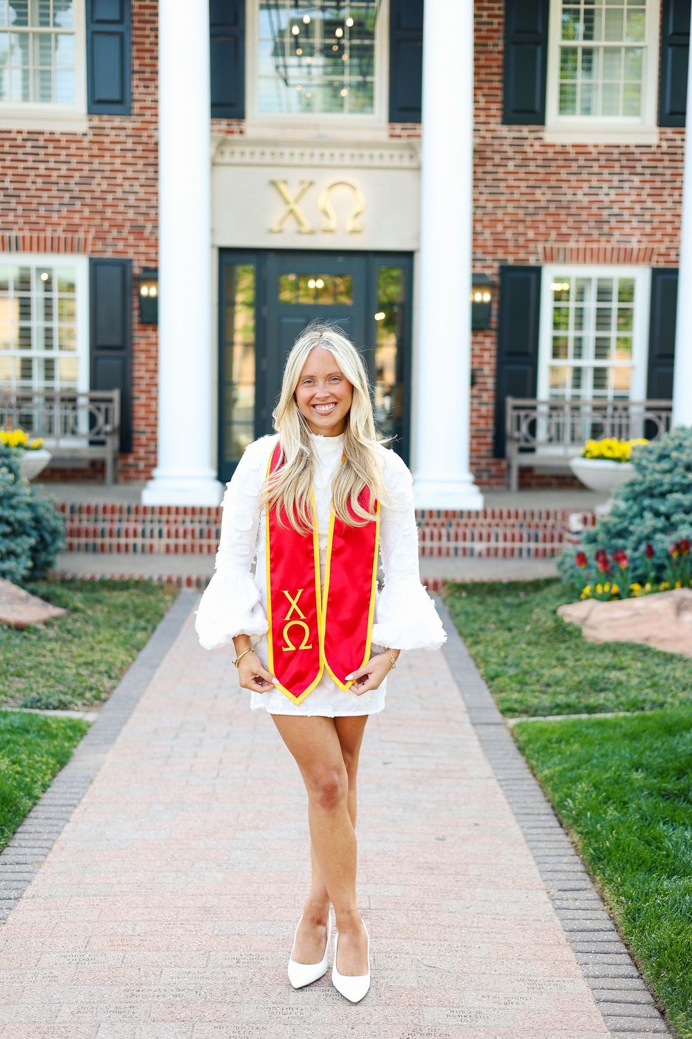 College student in red and white sorority letters poses on brick walkway in front of traditional university building.