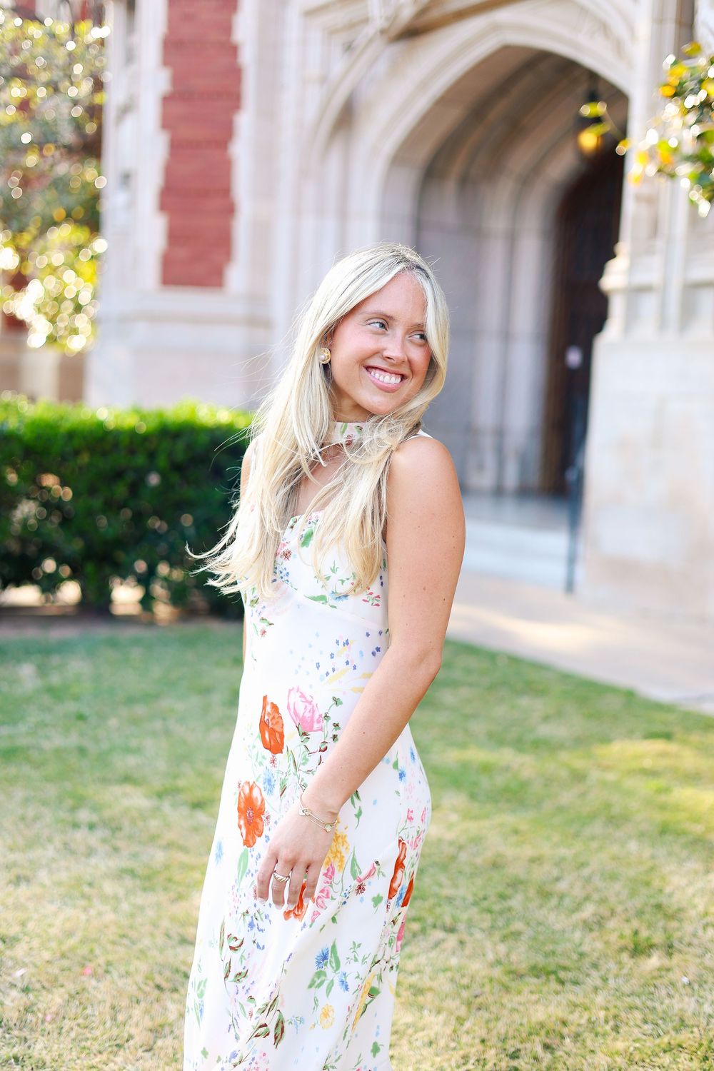 A person wearing a white floral dress smiles joyfully while standing on grass near a brick building.