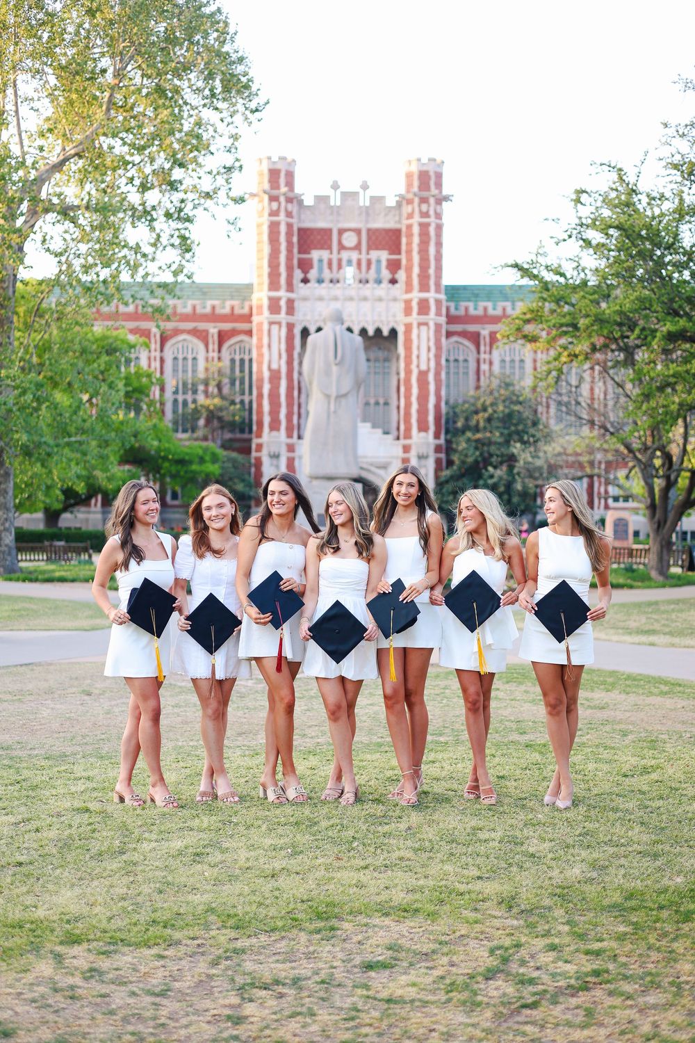 A group in white dresses poses with graduation caps on a college campus lawn with a historic brick building in background.