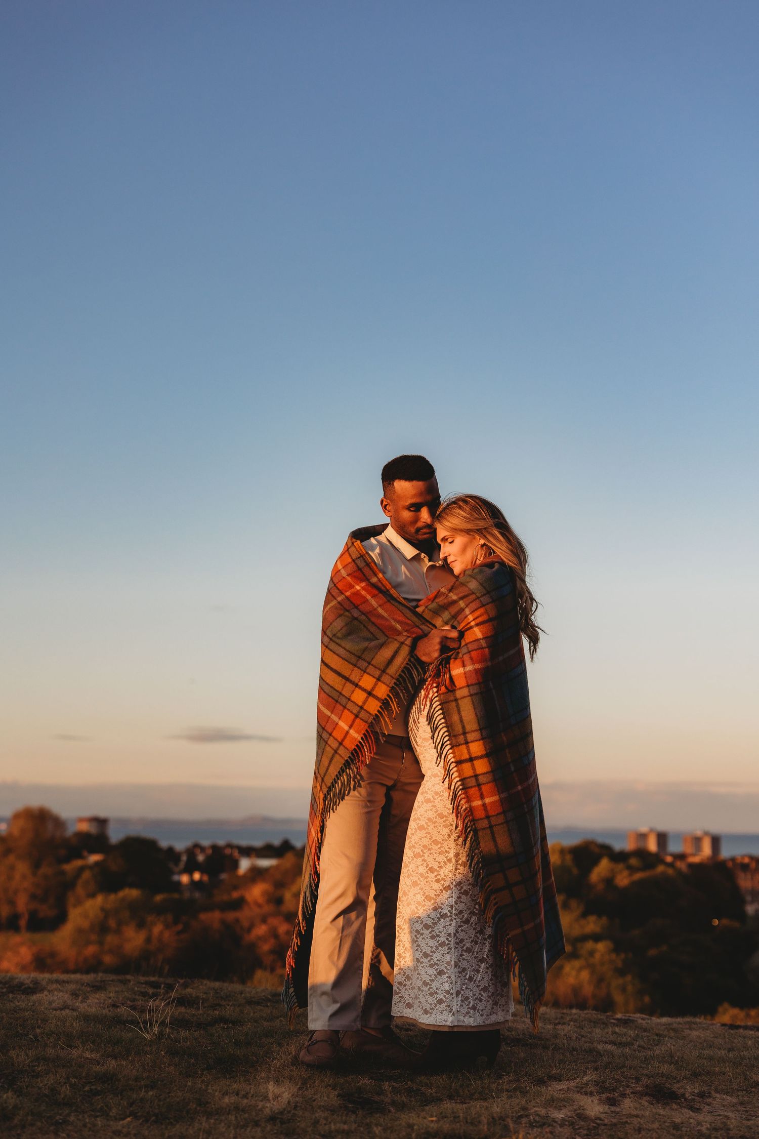 A couple wrapped in an orange plaid blanket stands together at sunset in a series of romantic outdoor photos.