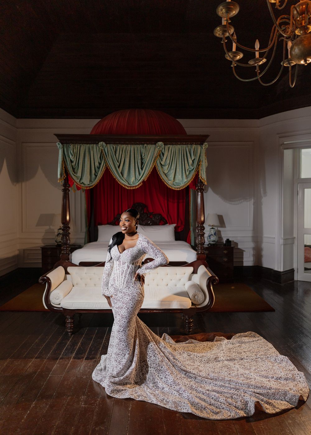 A flowing white lace gown with a dramatic train is showcased in an elegant room with dark hardwood floors and chandeliers.