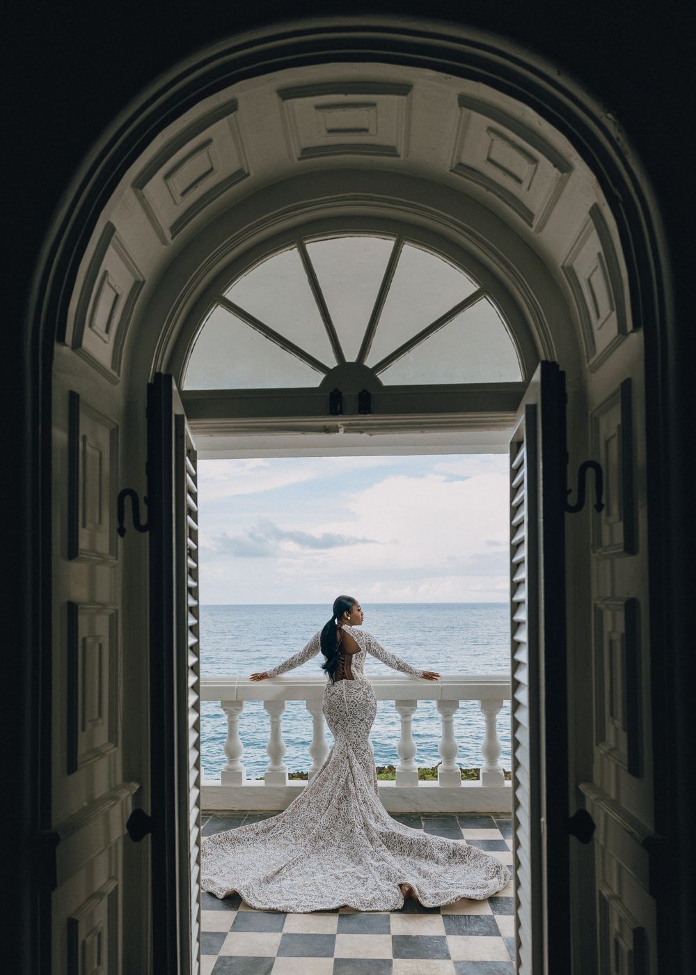 Elegant silhouette framed by an ornate archway overlooking the ocean from a balcony with checkered floor.