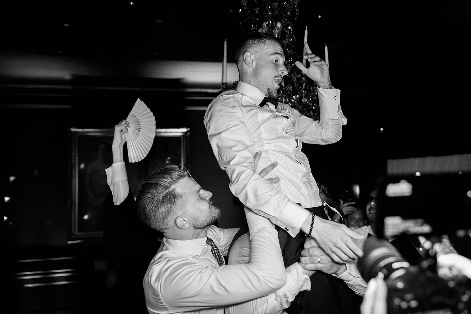 A black and white photo shows a person being lifted up during what appears to be a celebratory moment at an indoor event.