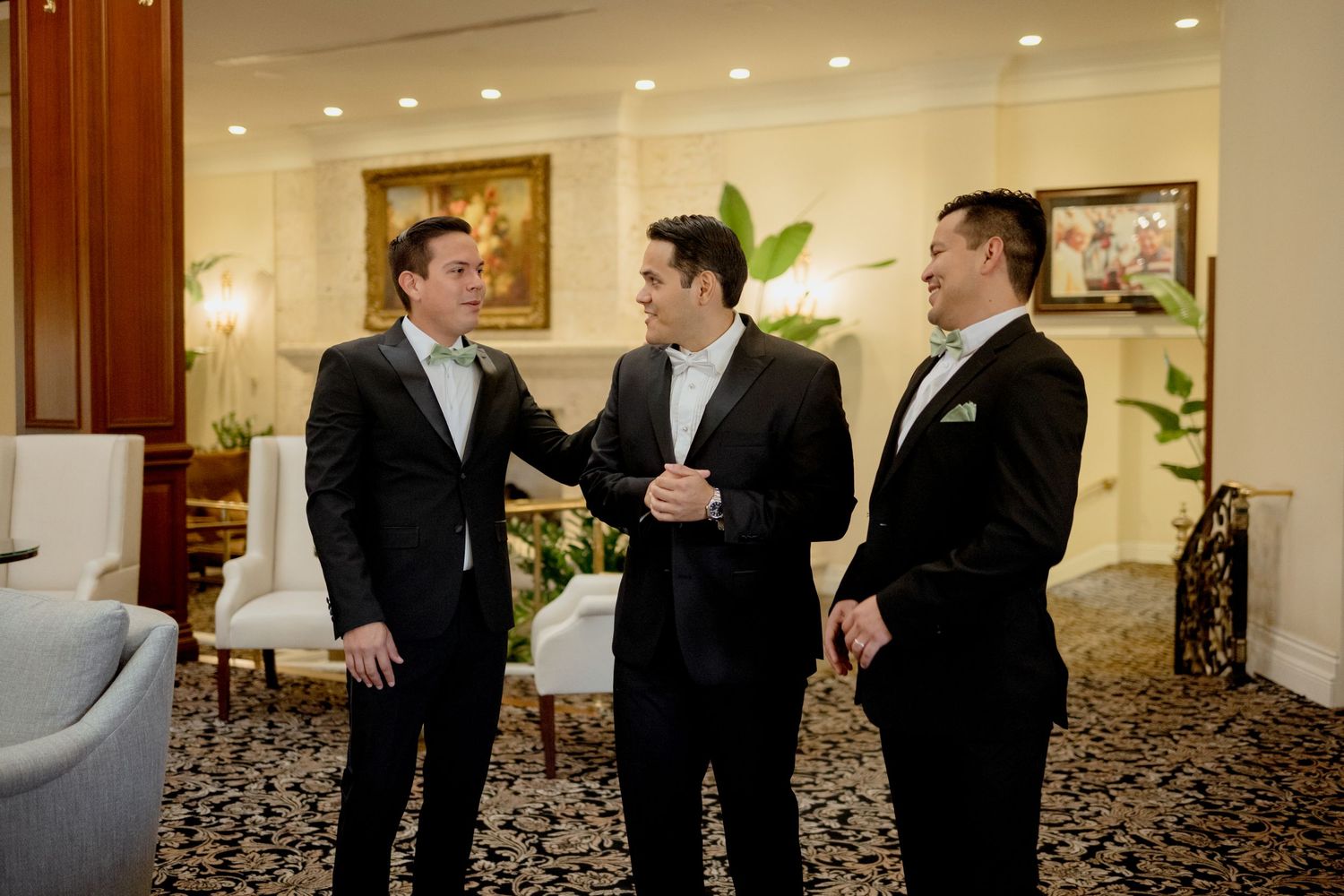A group in formal black suits and ties gather in conversation at an elegant indoor venue with carpeted floors and recessed lighting.