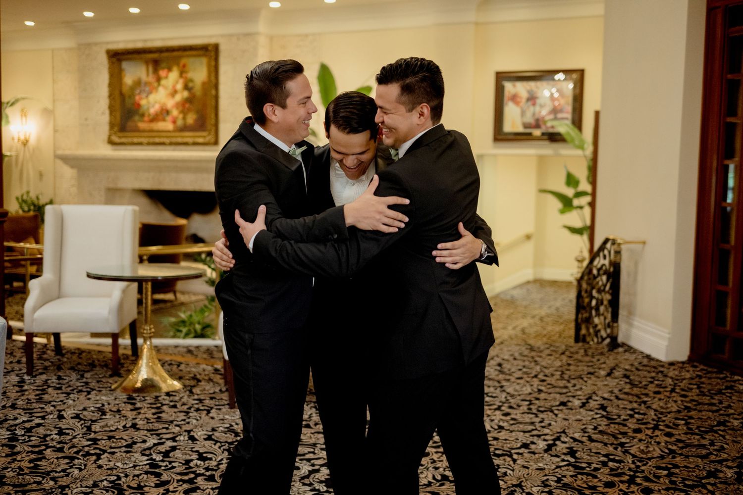 Three people in formal black suits embrace in a warm group hug at an elegant venue with floral artwork and carpeting.
