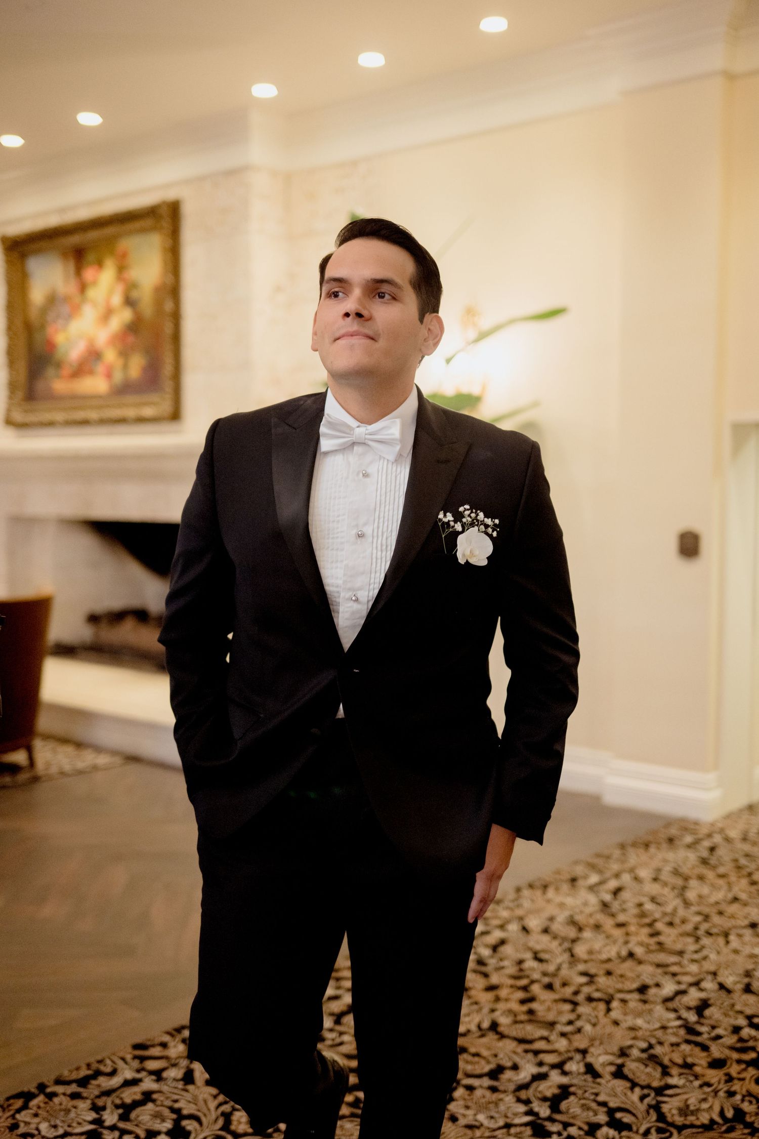 A person in a black tuxedo and white bow tie stands in an elegant room with patterned carpet and recessed lighting.