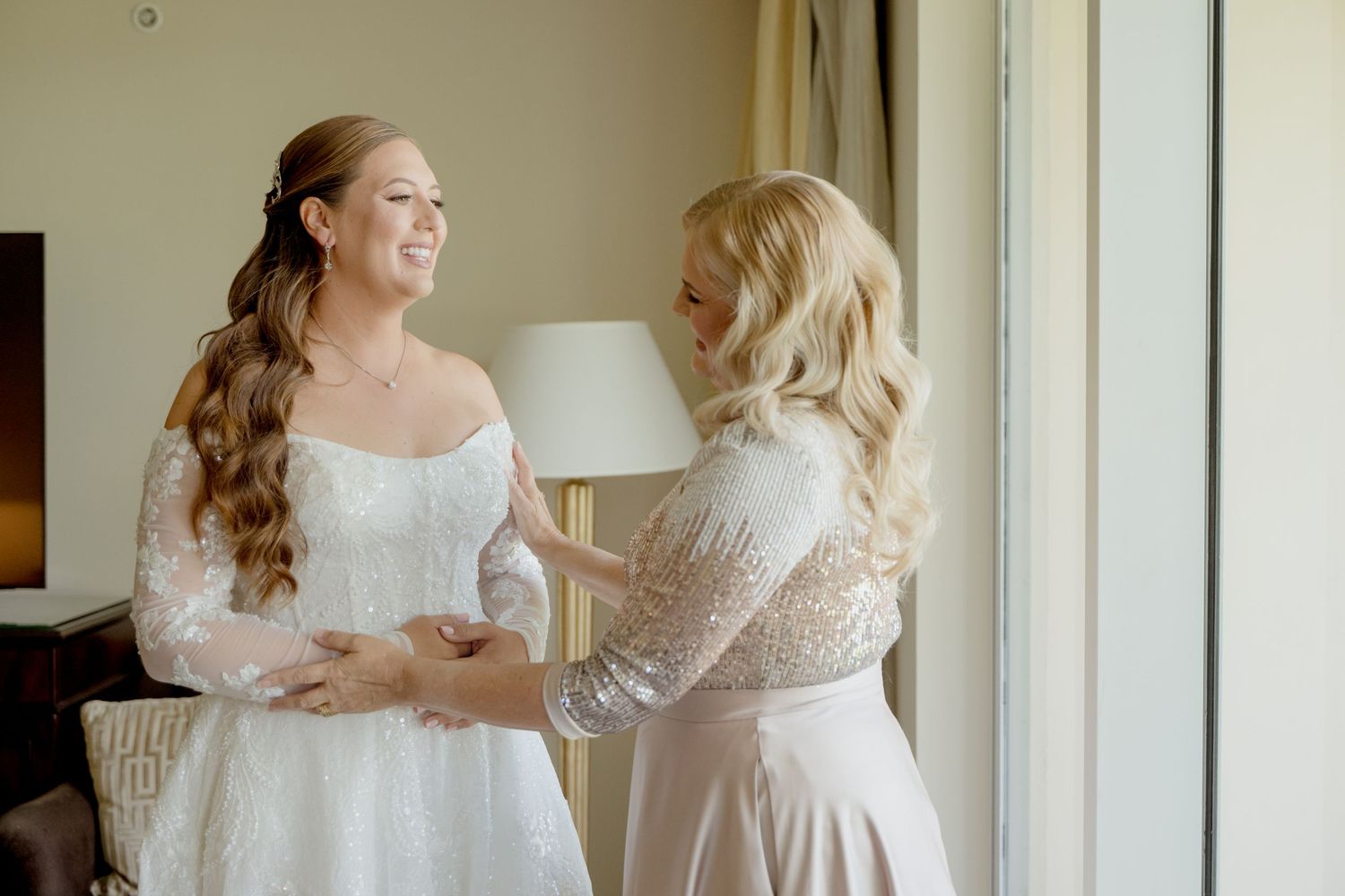 Two people in white lace dresses share a joyful moment together near large windows in a bright room.